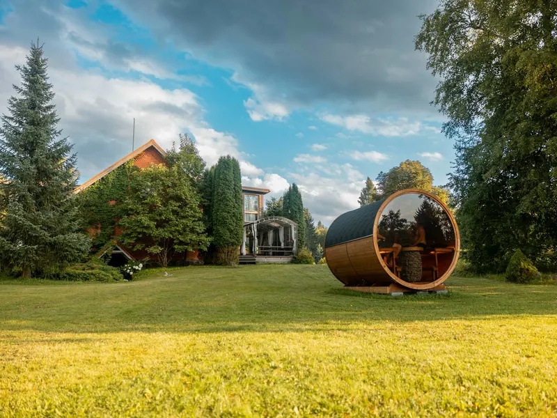 Wooden barrel sauna on a backyard lawn beside a house and tall trees
