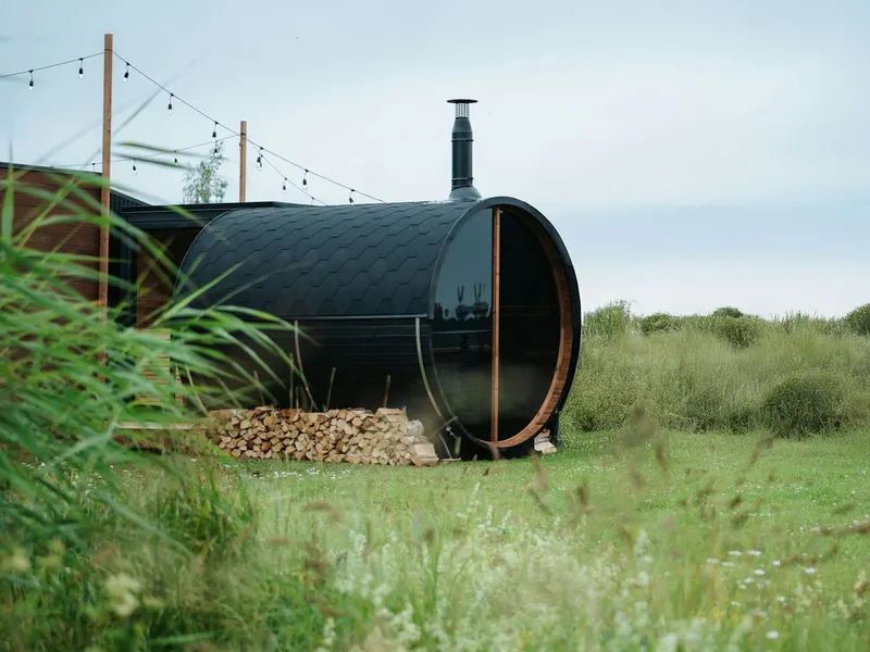 Black barrel sauna with a chimney and a stack of firewood in a grassy field