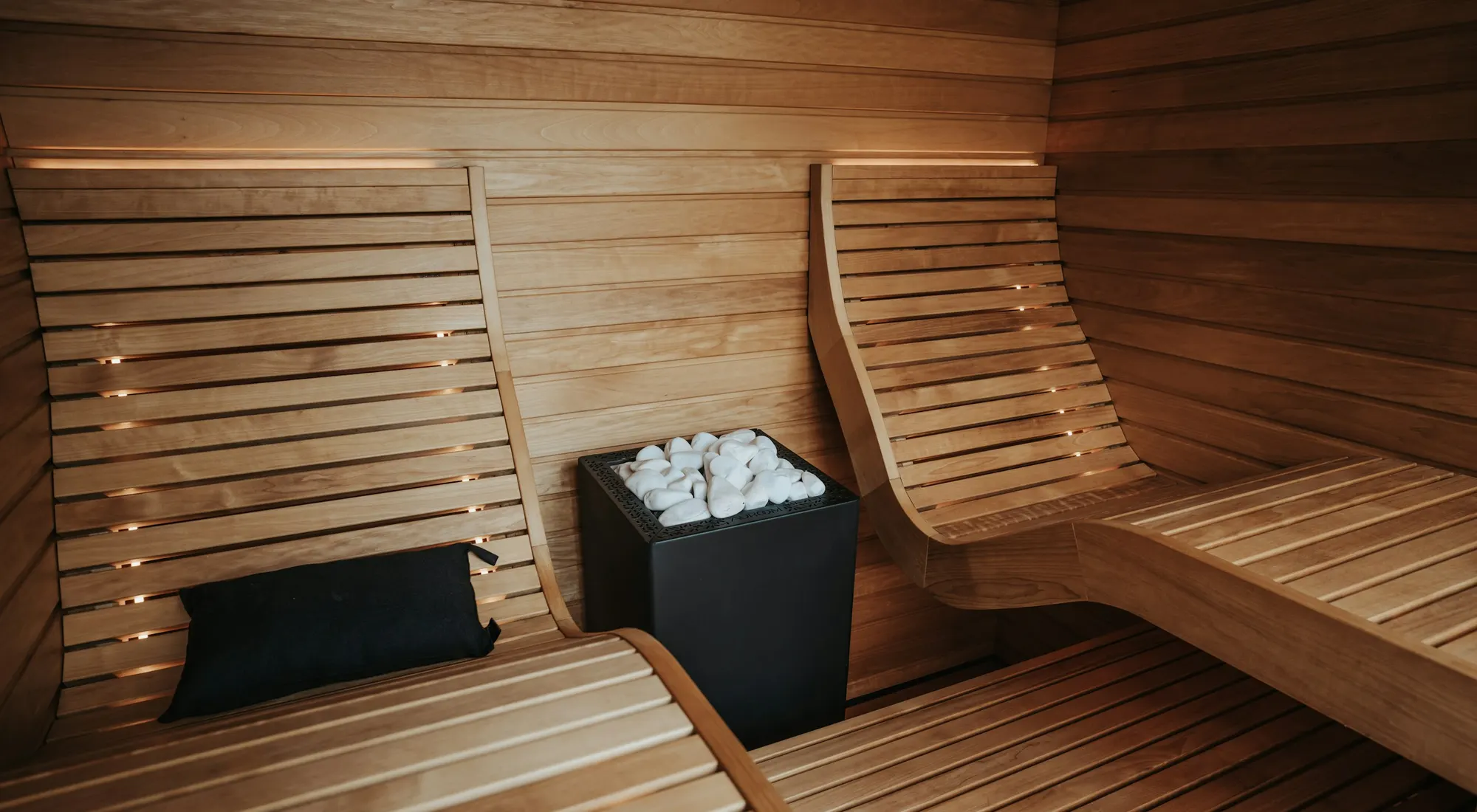 Wooden sauna with curved lounge benches and soft lighting around a black stone heater